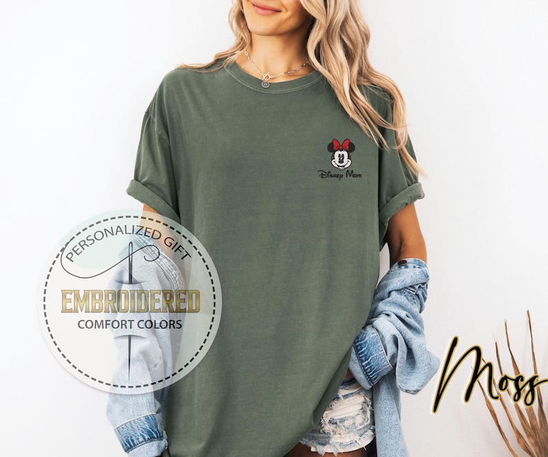 May include: A moss green t-shirt with rolled-up sleeves. The shirt features an embroidered Minnie Mouse design with a red bow and the text "Disney Mom." A circular logo reads "Personalized Gift" and "Embroidered Comfort Colors."