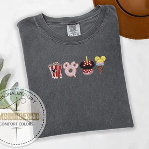 May include: A gray t-shirt with an embroidered design of Mickey Mouse themed snacks. The snacks include popcorn, a donut, a Mickey Mouse ice cream cone, and a Mickey Mouse ear ice cream. The text "COMFORT COLORS" is visible on the shirt.