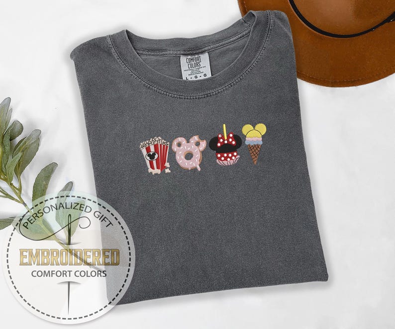 May include: A gray t-shirt with an embroidered design of Mickey Mouse themed snacks. The snacks include popcorn, a donut, a Mickey Mouse ice cream cone, and a Mickey Mouse ear ice cream. The text "COMFORT COLORS" is visible on the shirt.