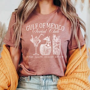 Puede incluir: Una camiseta rosa con el texto "Gulf of Mexico Social Club" y un gr&aacute;fico de cuatro c&oacute;cteles. El texto debajo del gr&aacute;fico dice "Stay Salty, Resist Often."