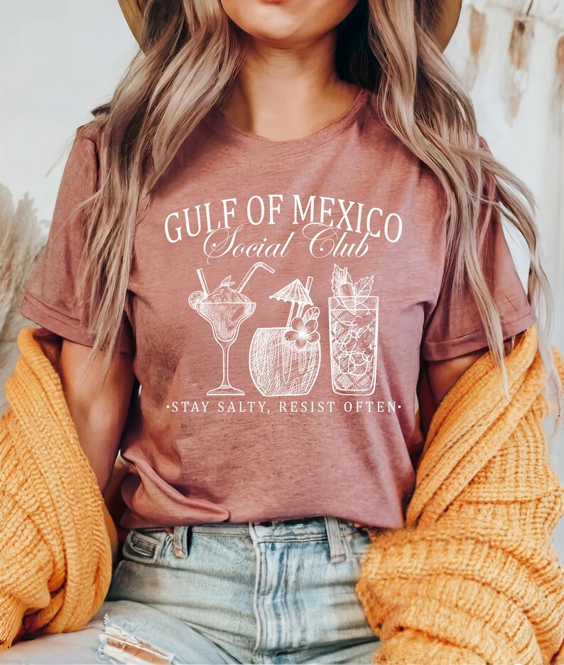 Puede incluir: Una camiseta rosa con el texto "Gulf of Mexico Social Club" y un gr&aacute;fico de cuatro c&oacute;cteles. El texto debajo del gr&aacute;fico dice "Stay Salty, Resist Often."