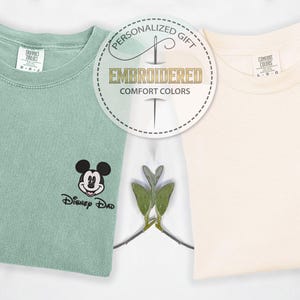 May include: Two embroidered Comfort Colors t-shirts, one mint green with a Mickey Mouse design and the text "Disney Dad", and one cream-colored with a Minnie Mouse design and the text "Disney Mom". A circular emblem reads "Personalized Gift, Embroidered, Comfort Colors".