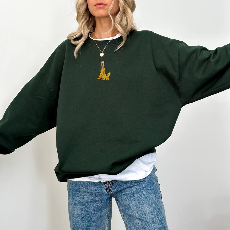 May include: Dark green sweatshirt with a small embroidered Pluto design in yellow and brown. The sweatshirt has long sleeves and a crew neck. A white t-shirt is visible underneath. The model is wearing blue jeans.
