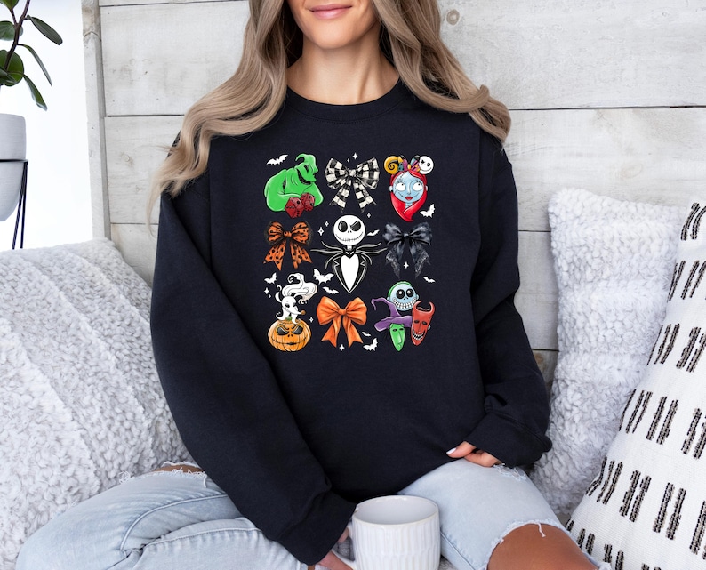 May include: Black sweatshirt with a colourful collage of Halloween-themed character bows. The design includes images of Jack Skellington, Sally, Oogie Boogie, and other characters from The Nightmare Before Christmas. The bows are in various colours and styles.