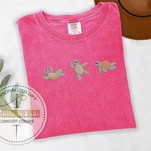 May include: A hot pink Comfort Colors t-shirt with three embroidered sea turtles in various poses. The shirt has a crew neck and short sleeves. The text "EMBROIDERED COMFORT COLORS" is visible on a circular sticker.