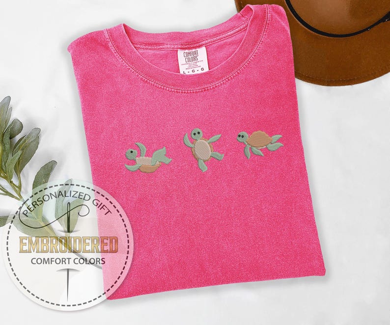 May include: A hot pink Comfort Colors t-shirt with three embroidered sea turtles in various poses. The shirt has a crew neck and short sleeves. The text "EMBROIDERED COMFORT COLORS" is visible on a circular sticker.