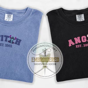 Puede incluir: Dos camisetas, una azul y otra negra, con dise&ntilde;os bordados. La camiseta azul tiene un personaje Stitch azul con el texto "STITCH EST. 2002". La camiseta negra tiene un personaje Angel rosa con el texto "ANGIL EST. 2002".