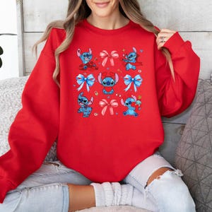 K&ouml;nnte beinhalten: Roter Pullover mit einem sich wiederholenden Muster aus blauen Stitch-Figuren, die Schleifen tragen. Stitch ist eine Zeichentrickfigur aus dem Disney-Film Lilo & Stitch.