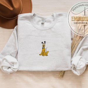 May include: Light gray sweatshirt with an embroidered yellow cartoon dog. The dog has black ears and a black nose. The sweatshirt has a crew neck and long sleeves. A brown hat is in the background.