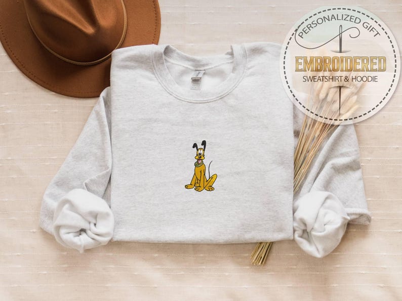 May include: Light gray sweatshirt with an embroidered yellow cartoon dog. The dog has black ears and a black nose. The sweatshirt has a crew neck and long sleeves. A brown hat is in the background.