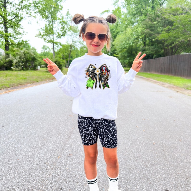 May include: A child wearing a white long-sleeved sweatshirt with a colorful graphic featuring characters. The child is also wearing black biker shorts with a white smiley face pattern and white socks. The child is wearing sunglasses and making a peace sign.