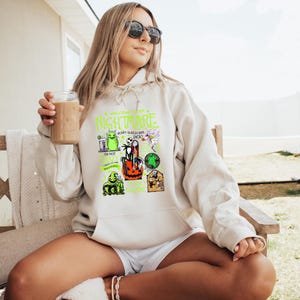 May include: A cream-coloured hoodie with a graphic featuring characters and text from "The Nightmare Before Christmas." The design includes images of Jack Skellington, Oogie Boogie, and other elements in green, orange, and black. The hoodie has a drawstring hood.
