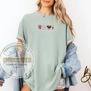 May include: A light green t-shirt with embroidered images of popcorn, a Mickey Mouse head, a pig, and an ice cream cone.