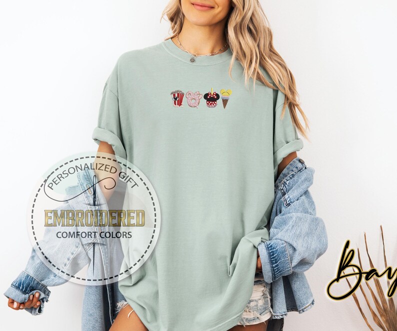 May include: A light green t-shirt with embroidered images of popcorn, a Mickey Mouse head, a pig, and an ice cream cone.