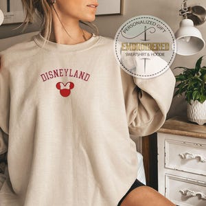 May include: A sand-colored sweatshirt with the text "DISNEYLAND" in red and a red embroidered Mickey Mouse head with a bow. The sweatshirt is personalized with the text "Personalized Gift Embroidered Sweatshirt & Hoodie".