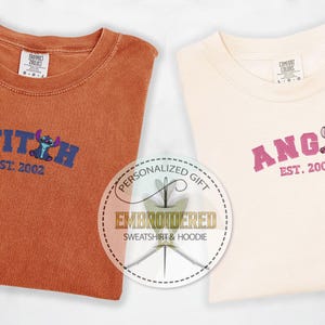 Puede incluir: Dos camisetas con dise&ntilde;os bordados. La camiseta naranja tiene "STITCH EST. 2002" en azul con un personaje Stitch azul. La camiseta blanca tiene "ANGEL EST. 2002" en rosa con un personaje Stitch rosa.