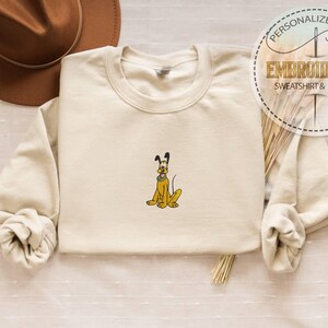 May include: Cream-colored sweatshirt with an embroidered Pluto design. The cartoon dog is yellow with black ears and a green collar. The sweatshirt has long sleeves and the text "PERSONALIZED GIFT EMBROIDERED SWEATSHIRT & HOODIE" in a circular design.