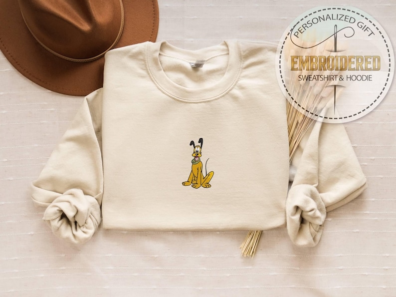 May include: Cream-colored sweatshirt with an embroidered Pluto design. The cartoon dog is yellow with black ears and a green collar. The sweatshirt has long sleeves and the text "PERSONALIZED GIFT EMBROIDERED SWEATSHIRT & HOODIE" in a circular design.