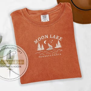 Pu&ograve; includere: Una maglietta color arancio bruciato con un disegno ricamato bianco che dice "Moon Lake Pennsylvania" con un lupo che ulula alla luna e pini.