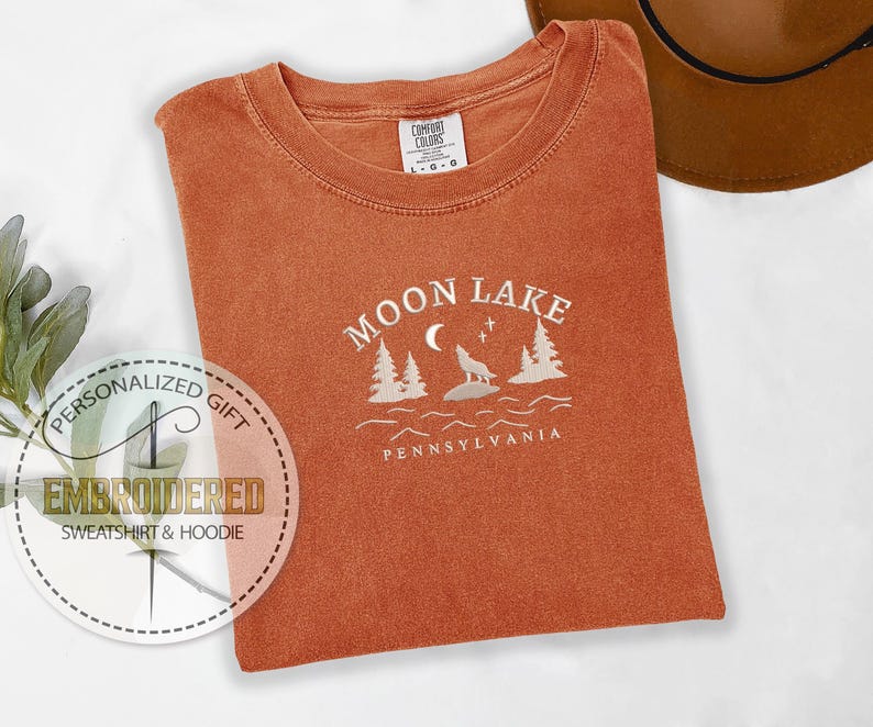 Pu&ograve; includere: Una maglietta color arancio bruciato con un disegno ricamato bianco che dice "Moon Lake Pennsylvania" con un lupo che ulula alla luna e pini.