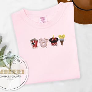 May include: Pink t-shirt with embroidered images of popcorn, a donut, a Mickey Mouse head, and an ice cream cone. The text "Personalized Gift" and "Embroidered Comfort Colors" is on the bottom left corner of the shirt.