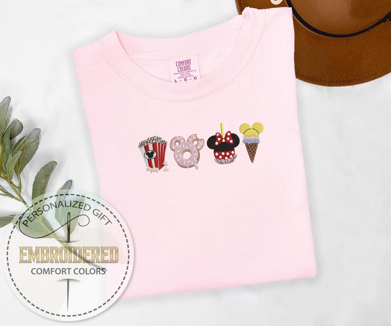 May include: Pink t-shirt with embroidered images of popcorn, a donut, a Mickey Mouse head, and an ice cream cone. The text "Personalized Gift" and "Embroidered Comfort Colors" is on the bottom left corner of the shirt.