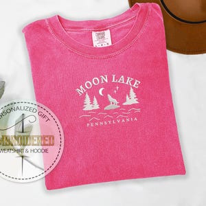 Pu&ograve; includere: Maglietta rosa con testo ricamato bianco che recita "Moon Lake Pennsylvania" con un lupo che ulula alla luna e pini.