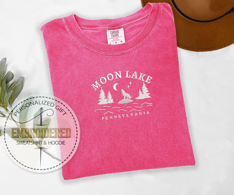 Pu&ograve; includere: Maglietta rosa con testo ricamato bianco che recita "Moon Lake Pennsylvania" con un lupo che ulula alla luna e pini.