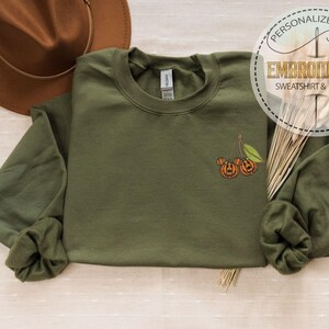 May include: An olive green sweatshirt with a crew neck and long sleeves. The sweatshirt features an embroidered design of two jack-o'-lanterns and a leaf. The text "PERSONALIZED GIFT EMBROIDERED SWEATSHIRT & HOODIE" is in a circle.
