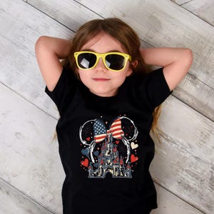 May include: A black t-shirt featuring a graphic of a castle, hearts, and a bow with American flag design. The child is wearing yellow sunglasses. The shirt design includes red, white, and blue colors.