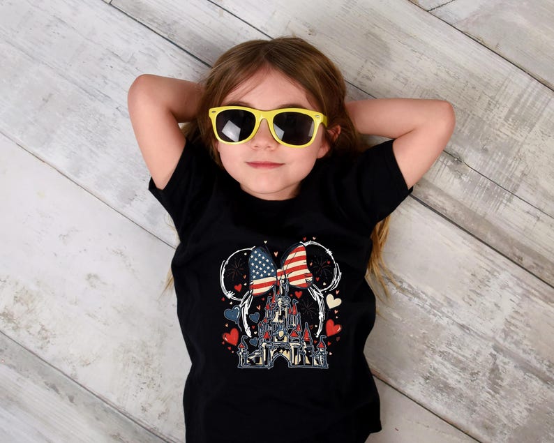 May include: A black t-shirt featuring a graphic of a castle, hearts, and a bow with American flag design. The child is wearing yellow sunglasses. The shirt design includes red, white, and blue colors.