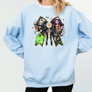 May include: Light blue sweatshirt featuring a large bow design with characters from The Nightmare Before Christmas. The bow is black with gold trim and a white daisy in the center. The sweatshirt has long sleeves and a crew neck.