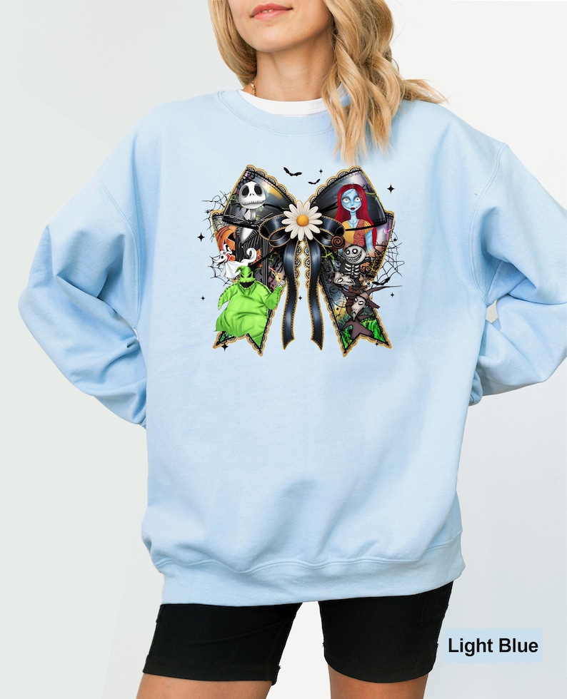 May include: Light blue sweatshirt featuring a large bow design with characters from The Nightmare Before Christmas. The bow is black with gold trim and a white daisy in the center. The sweatshirt has long sleeves and a crew neck.