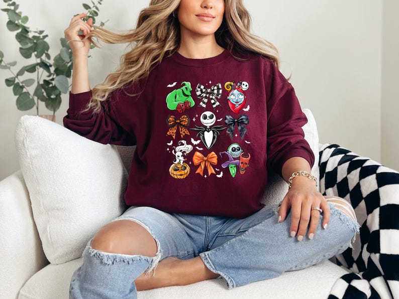 May include: Burgundy sweatshirt with a colourful graphic of characters and bows from The Nightmare Before Christmas. The design includes Jack Skellington, Sally, Oogie Boogie, and other iconic elements. The sweatshirt is a comfortable, casual style.