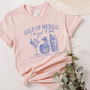 Puede incluir: Camiseta rosa con un gr&aacute;fico azul que dice "Gulf of Mexico Social Club" y presenta un dibujo de cuatro bebidas tropicales. El texto debajo del gr&aacute;fico dice "Stay Salty, Resist Often."
