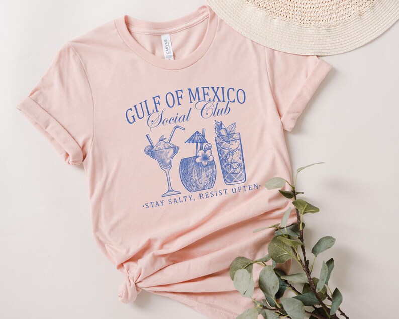 Puede incluir: Camiseta rosa con un gr&aacute;fico azul que dice "Gulf of Mexico Social Club" y presenta un dibujo de cuatro bebidas tropicales. El texto debajo del gr&aacute;fico dice "Stay Salty, Resist Often."