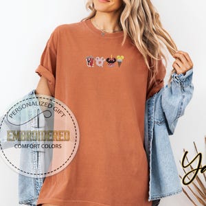 May include: A rust-colored t-shirt with an embroidered design of a popcorn bucket, Mickey Mouse head, ice cream cone, and a churro. The text "Personalized Gift" and "Embroidered Comfort Colors" is visible on a white circle.