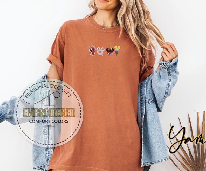 May include: A rust-colored t-shirt with an embroidered design of a popcorn bucket, Mickey Mouse head, ice cream cone, and a churro. The text "Personalized Gift" and "Embroidered Comfort Colors" is visible on a white circle.