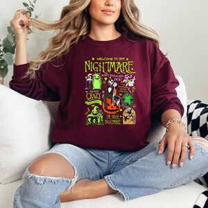 May include: Burgundy crew neck sweatshirt featuring a graphic from "The Nightmare Before Christmas." The design includes characters like Jack Skellington, Sally, and Oogie Boogie, with text that reads "Welcome to our Nightmare."
