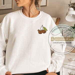 May include: White sweatshirt with a crew neck and long sleeves. The sweatshirt features an embroidered design of two orange pumpkins with green leaves. The text "PERSONALIZED GIFT EMBROIDERED SWEATSHIRT & HOODIE" is printed on a circular graphic.