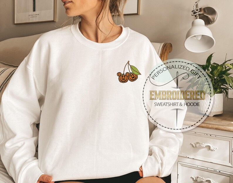 May include: White sweatshirt with a crew neck and long sleeves. The sweatshirt features an embroidered design of two orange pumpkins with green leaves. The text "PERSONALIZED GIFT EMBROIDERED SWEATSHIRT & HOODIE" is printed on a circular graphic.