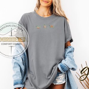 May include: A gray t-shirt with three embroidered sea turtles in shades of green, orange, and yellow. The shirt has a relaxed fit and is part of the Comfort Colors collection. The image also includes the text "Personalized Gift" and "Embroidered".