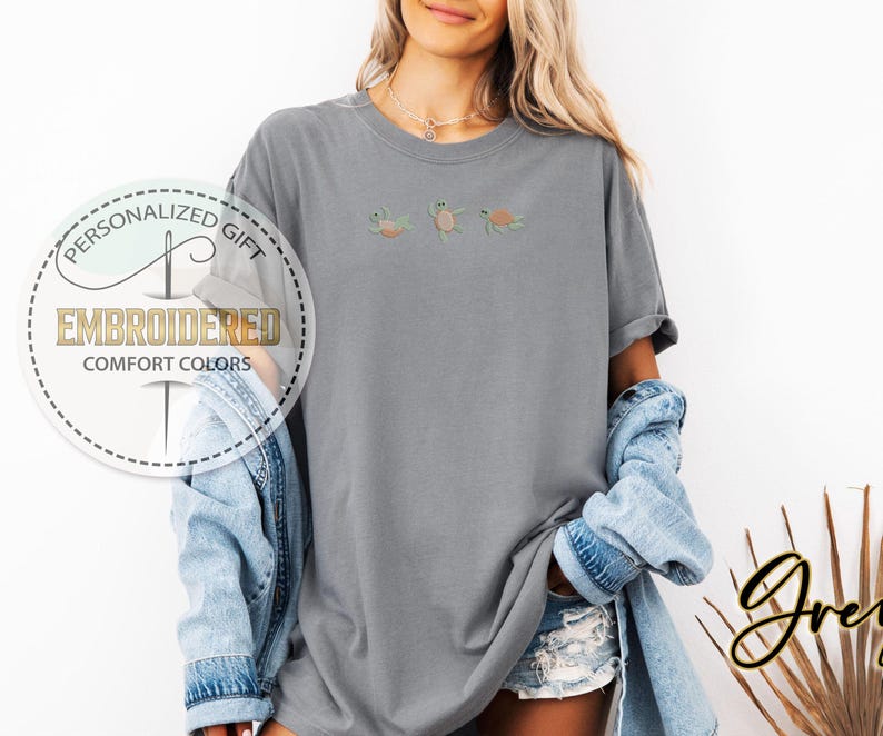 May include: A gray t-shirt with three embroidered sea turtles in shades of green, orange, and yellow. The shirt has a relaxed fit and is part of the Comfort Colors collection. The image also includes the text "Personalized Gift" and "Embroidered".
