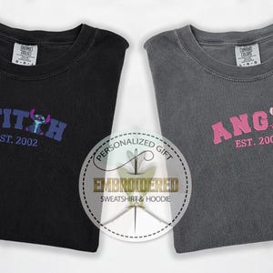 Puede incluir: Dos camisetas grises con dise&ntilde;os bordados. La camiseta de la izquierda tiene un dise&ntilde;o de Stitch azul y morado con el texto "STITCH EST. 2002". La camiseta de la derecha tiene un dise&ntilde;o de Angel rosa con el texto "ANGEL EST. 2002".