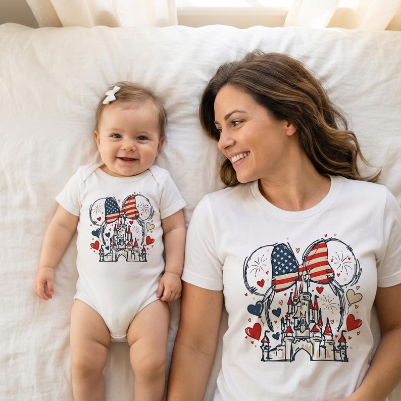 May include: White t-shirt and baby onesie featuring a patriotic design. The design includes a castle, hearts, and a bow with the American flag. The baby onesie is next to a matching t-shirt.