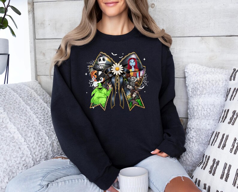 May include: Black crew neck sweatshirt featuring a large bow design with characters from The Nightmare Before Christmas. The bow is black with gold trim and a white daisy in the center. The characters include Jack Skellington, Sally, and Oogie Boogie.