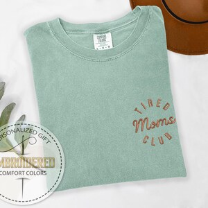 May include: A light green t-shirt with the text "TIRED MOMS CLUB" embroidered in orange on the front.