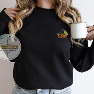 May include: Black sweatshirt with an embroidered design of two orange pumpkins with green leaves. The sweatshirt has a crew neck and long sleeves. A white mug is held in the right hand. The text "PERSONALIZED GIFT EMBROIDERED SWEATSHIRT & HOODIE" is on the left side.