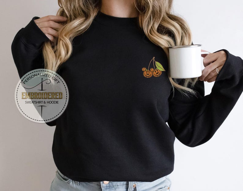 May include: Black sweatshirt with an embroidered design of two orange pumpkins with green leaves. The sweatshirt has a crew neck and long sleeves. A white mug is held in the right hand. The text "PERSONALIZED GIFT EMBROIDERED SWEATSHIRT & HOODIE" is on the left side.