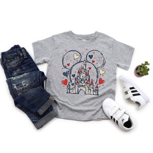 May include: A gray t-shirt featuring a castle design within a Mickey Mouse head outline, with red and blue hearts. The shirt is paired with blue jeans and white sneakers with black stripes. A small potted plant is also present.